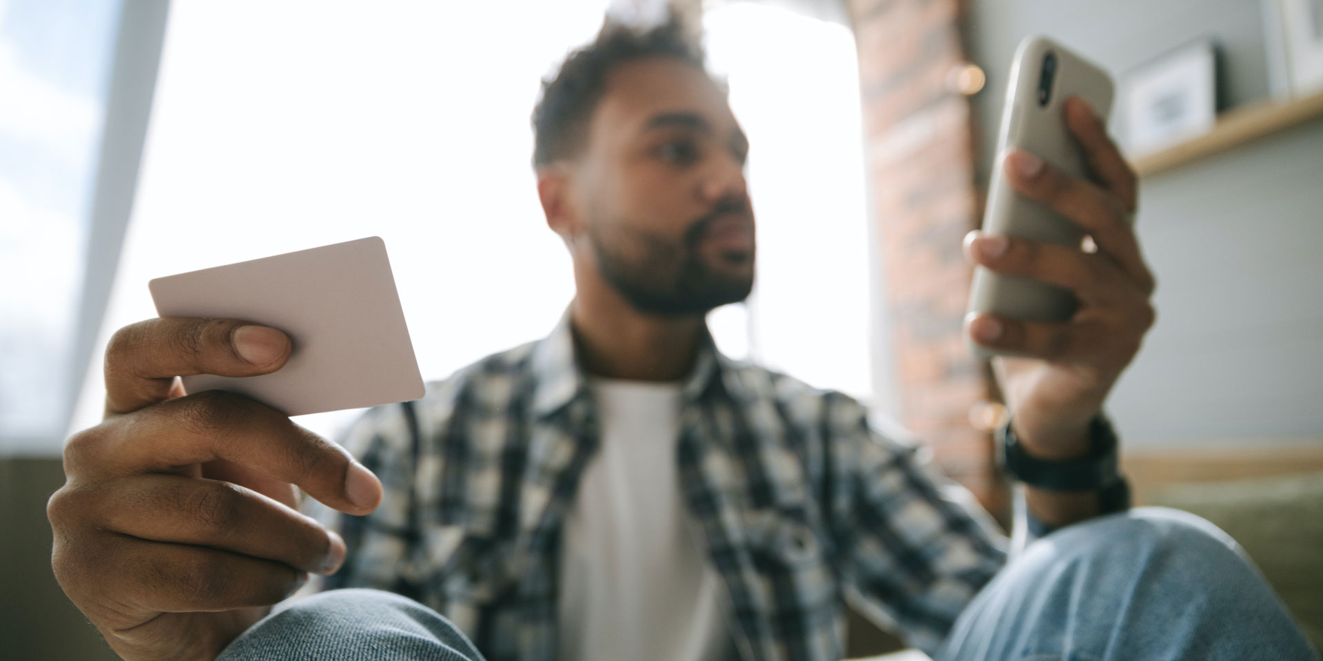 Young African man holding his bank card in one hand while holding and transacting on his cell phone in his other hand.